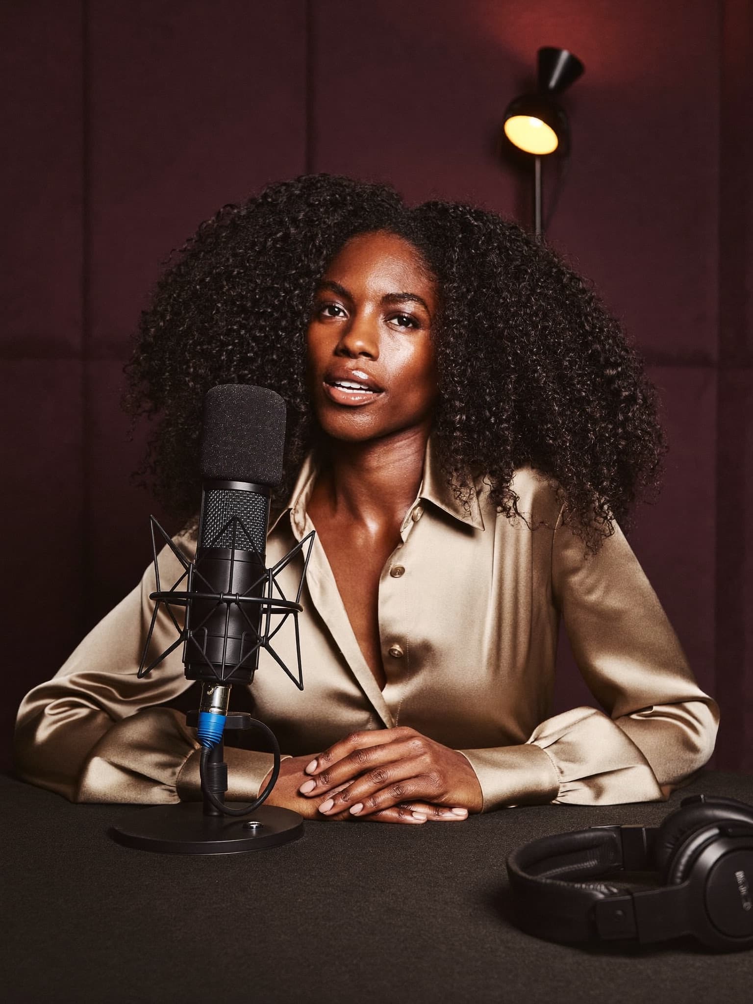 A confident woman in a champagne silk blouse speaks into a broadcast microphone in a warmly lit studio.