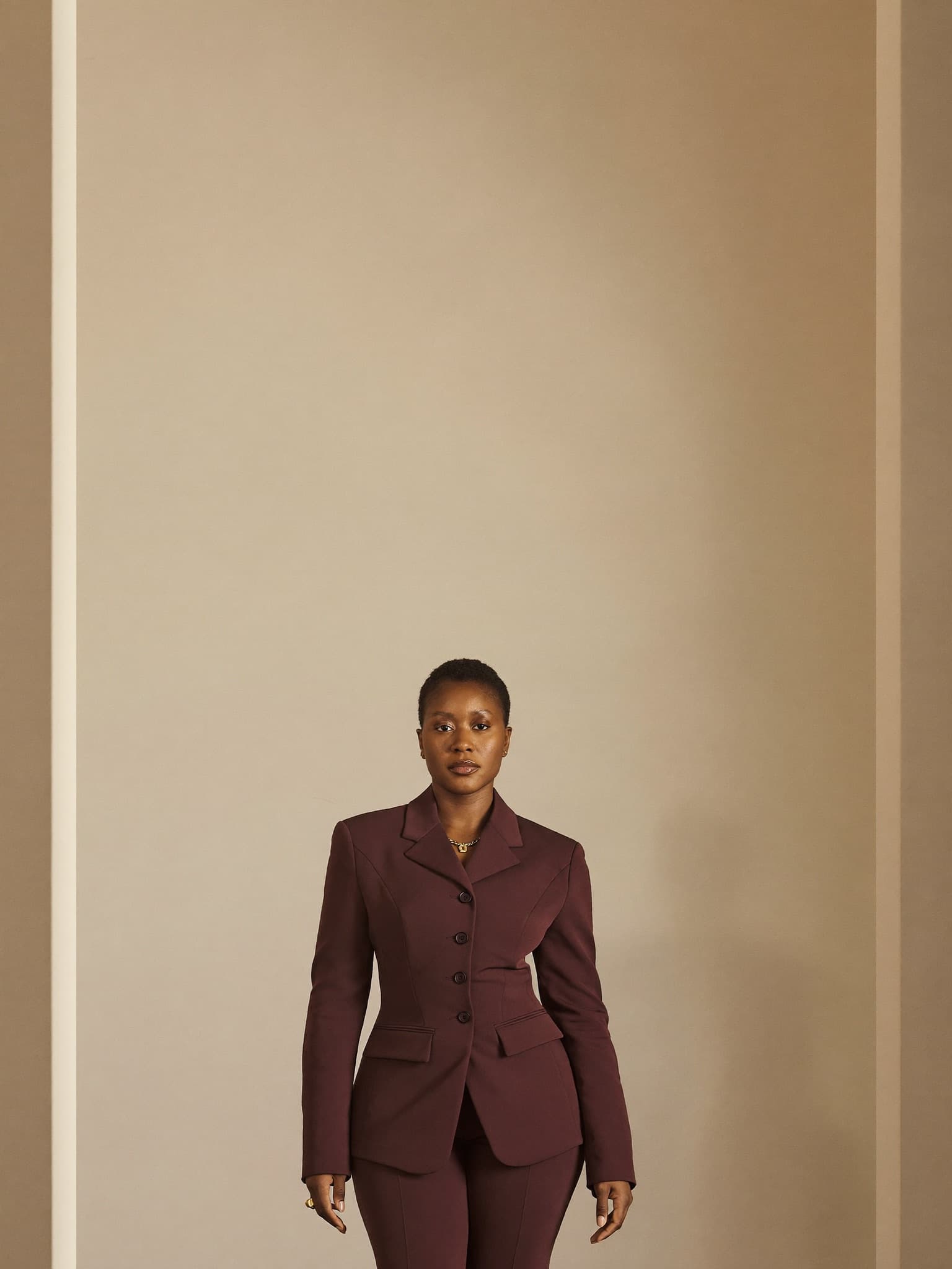 A poised woman in a tailored aubergine suit stands in a minimal ivory gallery, facing the camera with calm confidence.