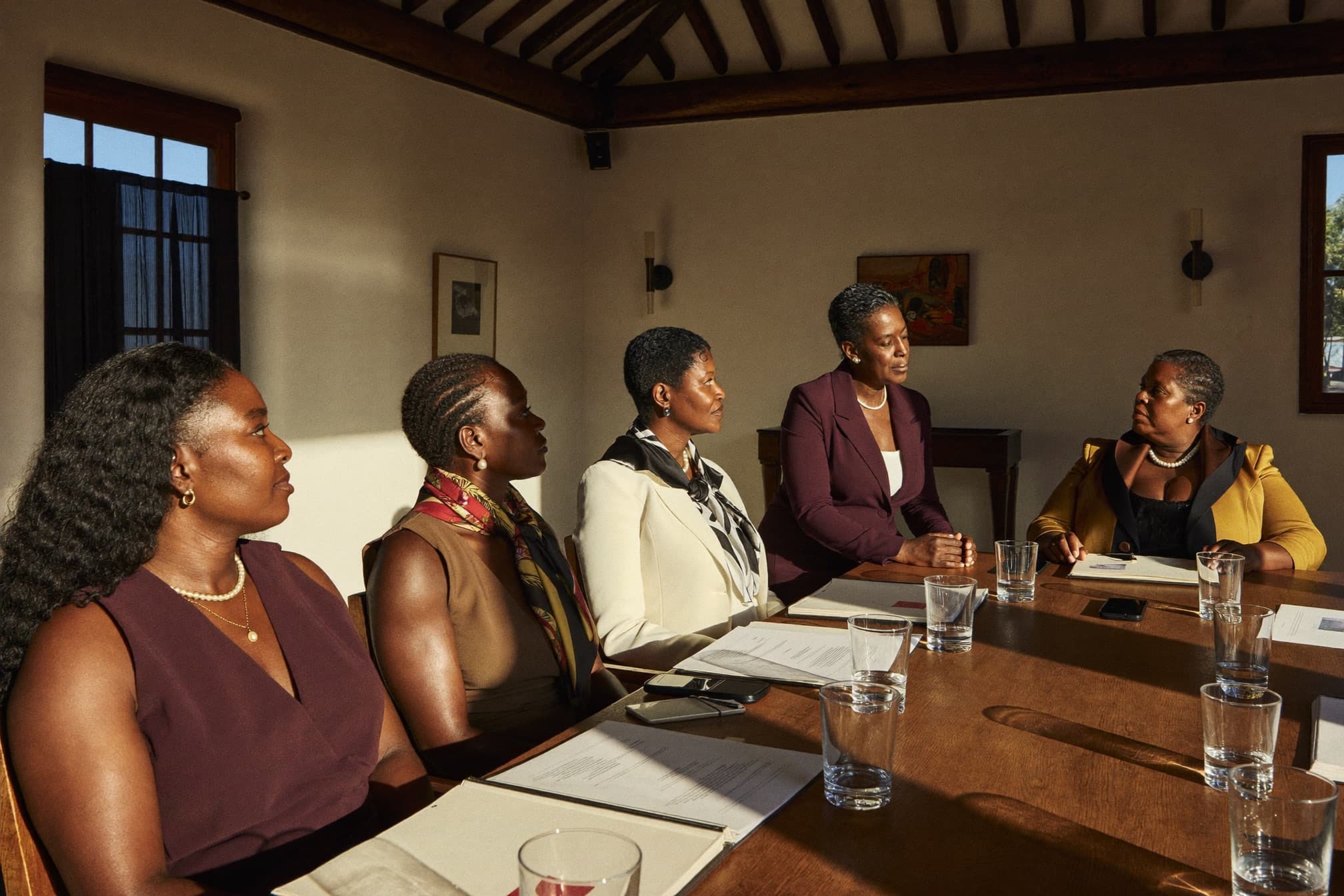 A group of Caribbean women in a sunlit workshop, one standing and speaking while the others listen with engagement.