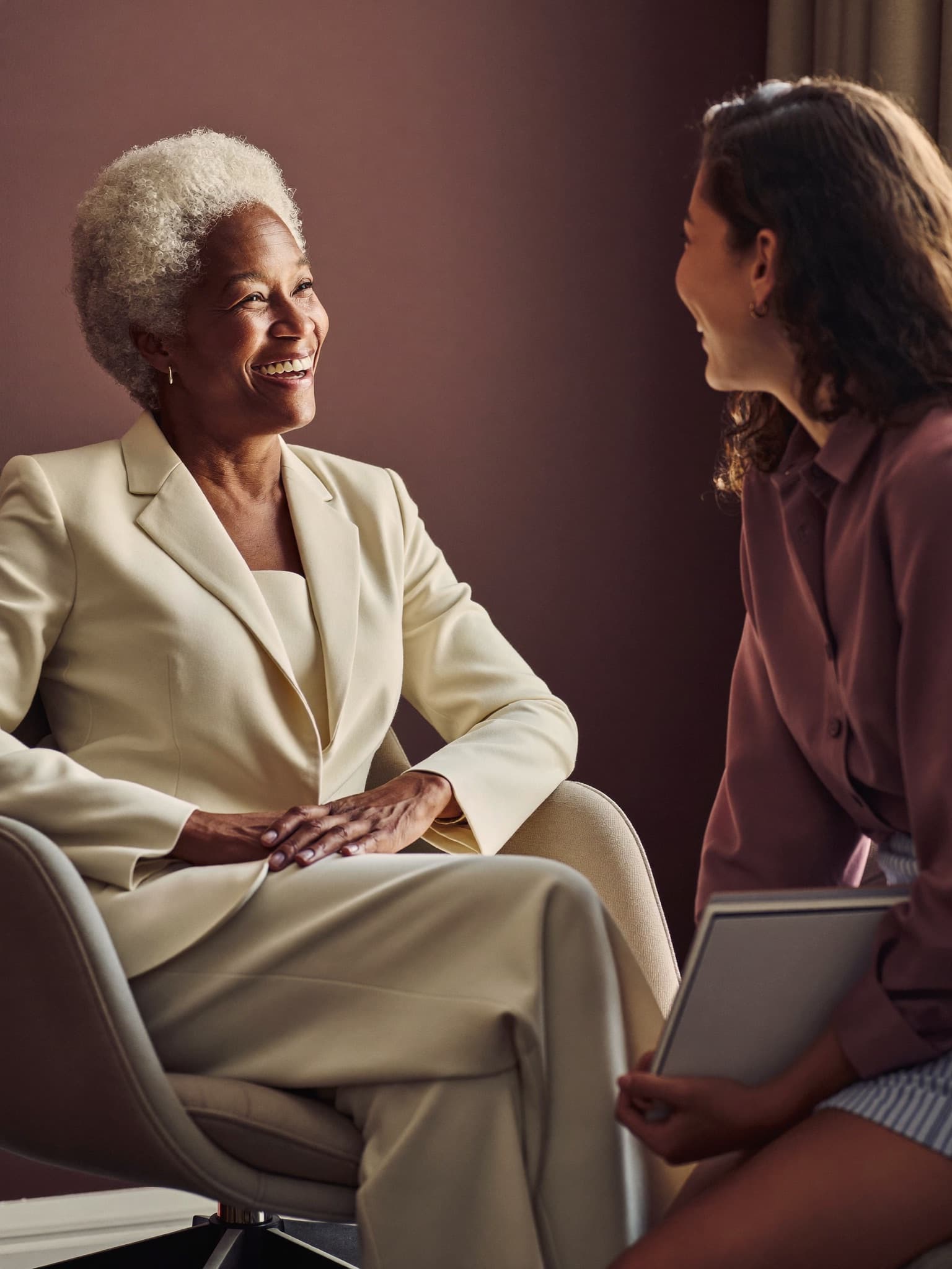 An accomplished older woman in an ivory suit shares a warm conversation with a younger woman holding a notebook.
