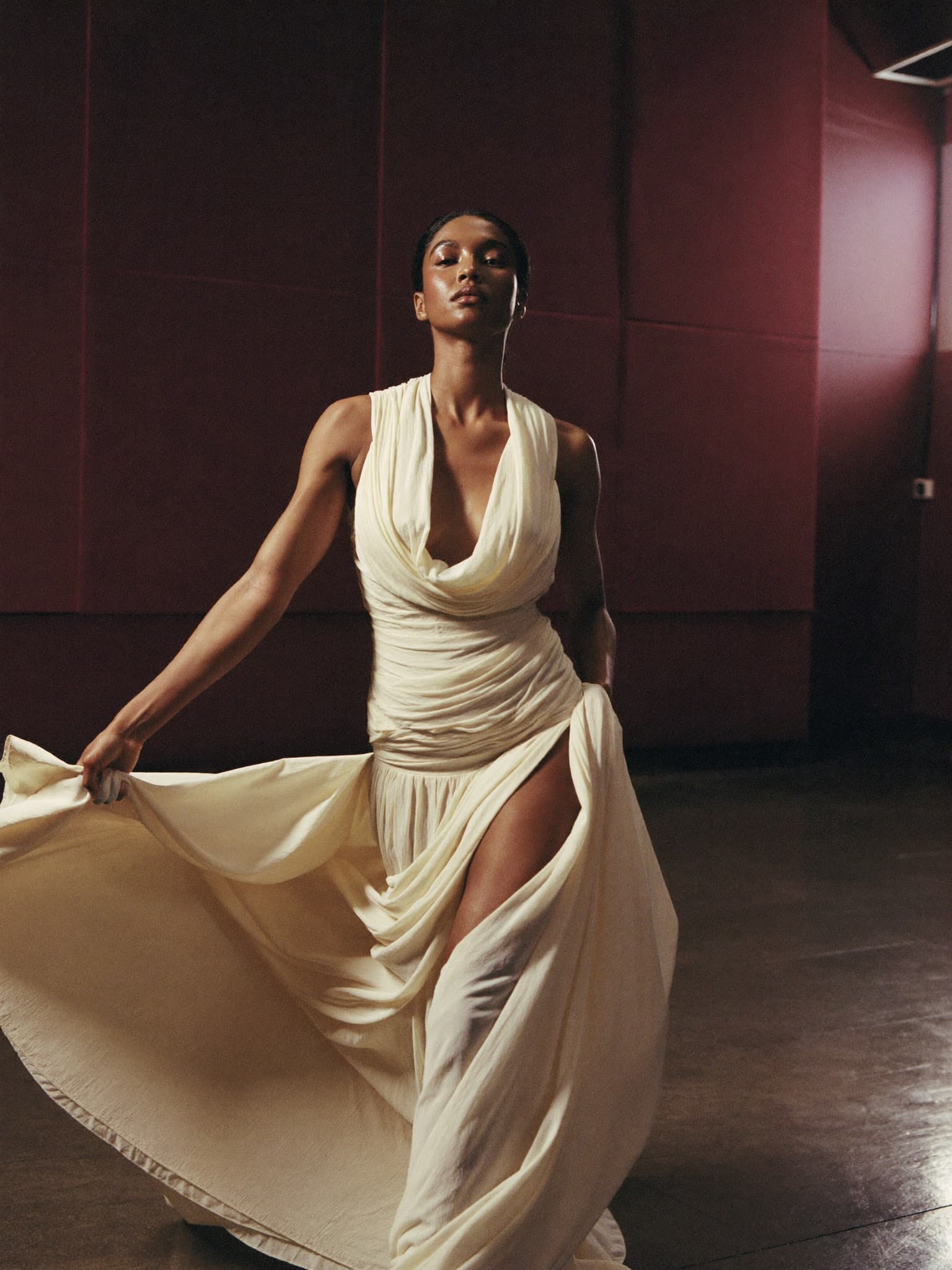 An elegant woman in a flowing ivory gown walks mid-stride through a rehearsal studio with deep plum walls.